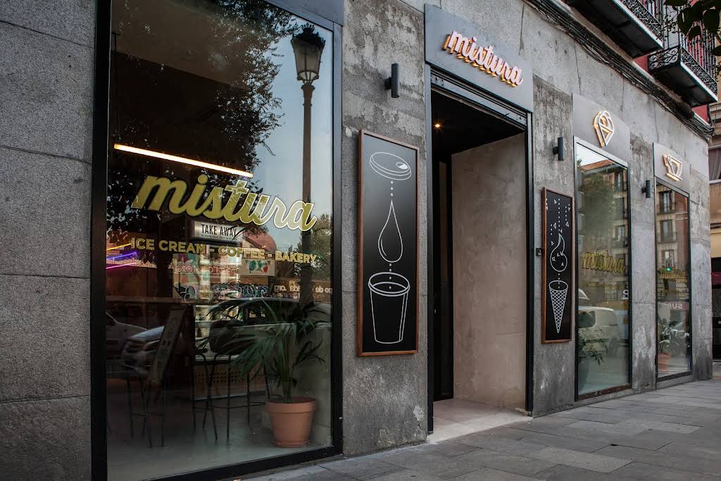Exterior de Mistura calle de Toledo con grandes ventanales, cartelería ilustrada y el rótulo dorado sobre fachada de piedra.