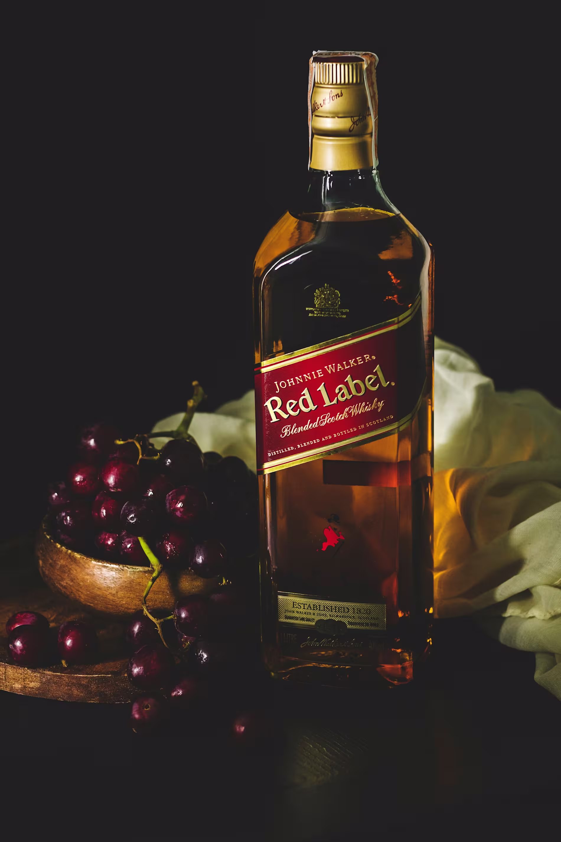 A bottle of Johnnie Walker Red Label whisky stands next to a wooden bowl filled with red grapes, set against a dark background with a cream-colored cloth.