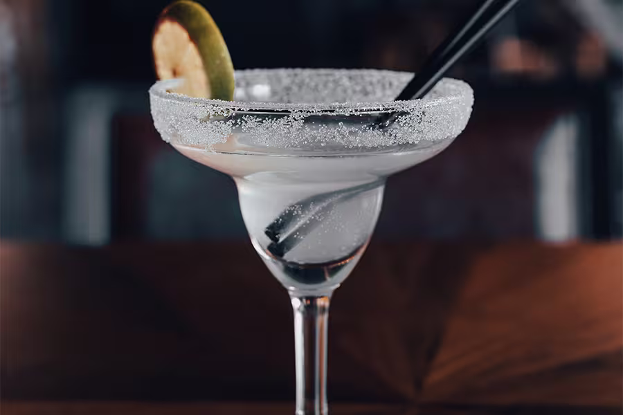 A margarita cocktail in a stemmed glass with a salted rim, garnished with a lime slice and served with two black straws.
