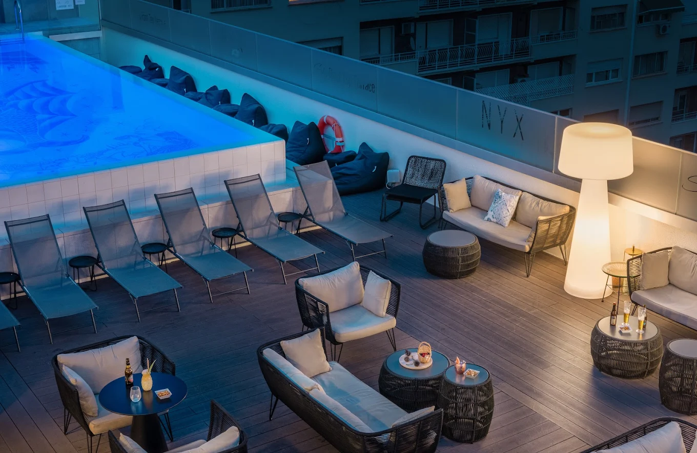 NYX Rooftop Terrace con piscina iluminada, tumbonas, sofás de exterior y mesas con copas en una elegante azotea urbana.