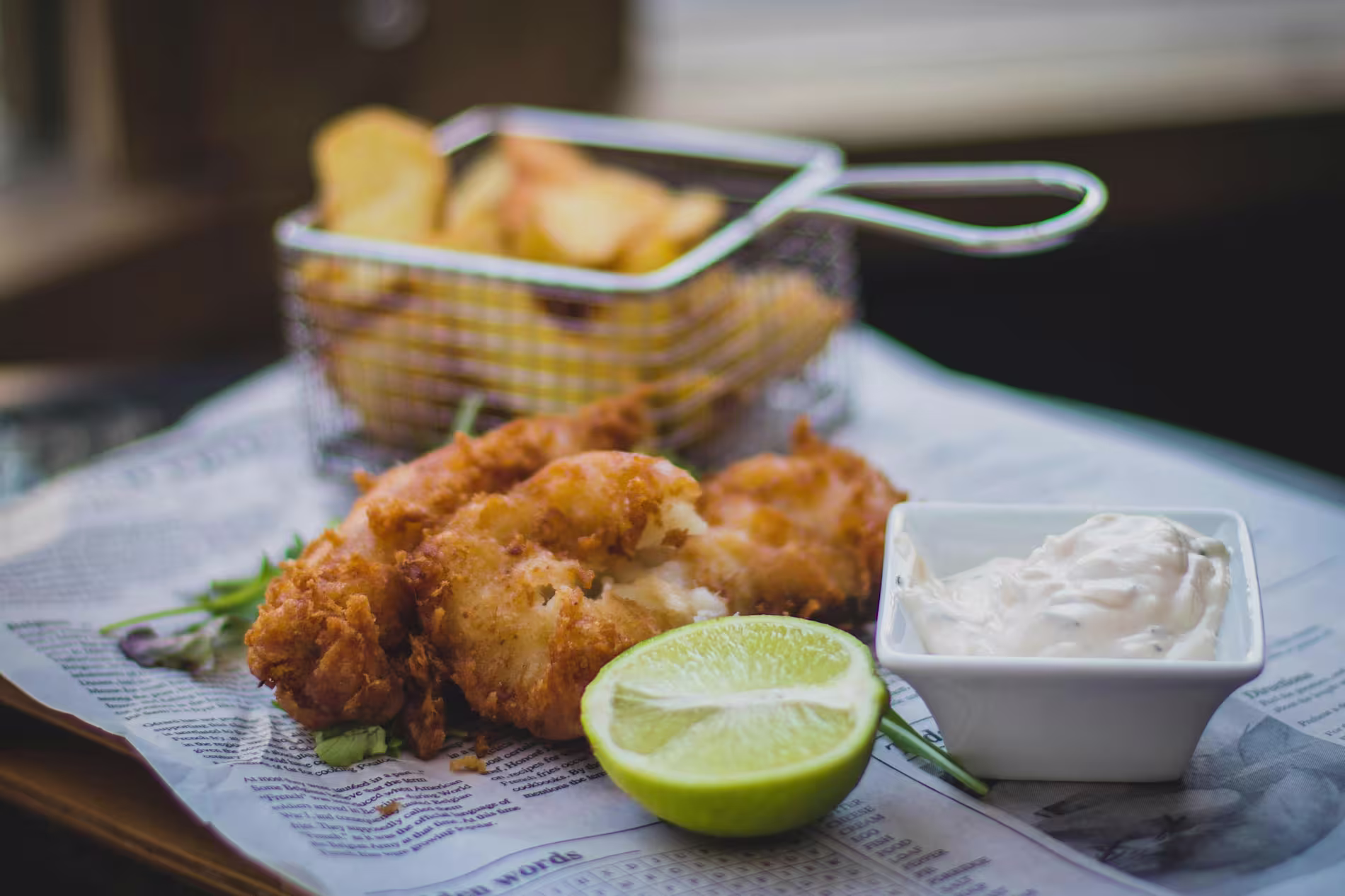A serving of battered fish with a lime wedge, a small dish of tartar sauce, and a basket of potato wedges on newspaper.