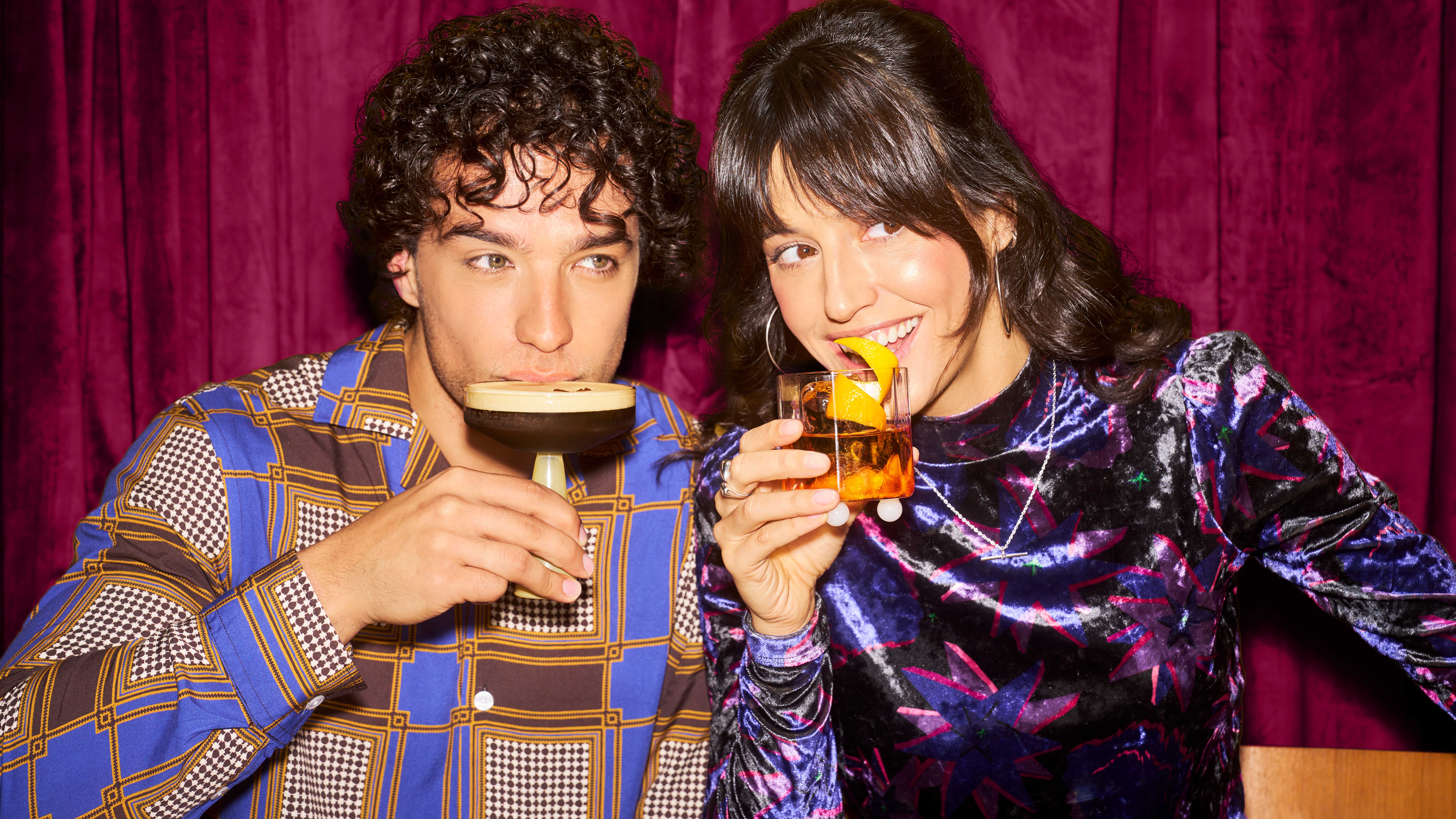 Un bărbat și o femeie în ținute colorate sorb cocktailuri în timp ce zâmbesc dincolo de cameră.