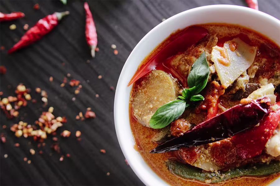A bowl of spicy red curry with vegetables, garnished with basil leaves and dried chili peppers, on a dark wooden surface scattered with chili flakes and whole chilies.