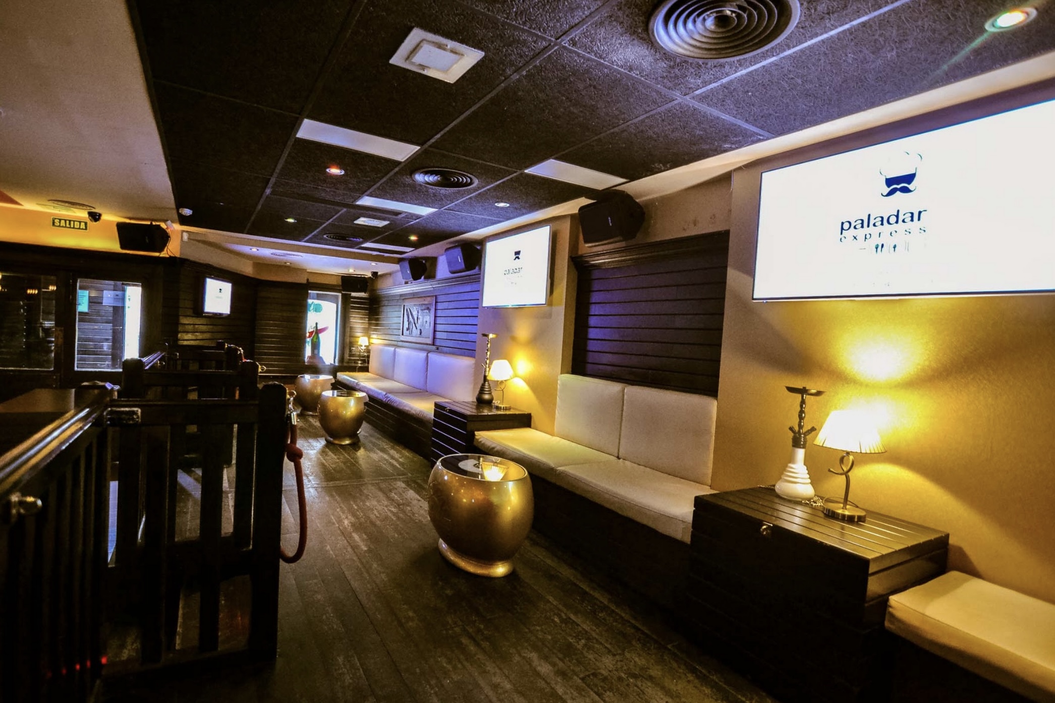 La imagen muestra el interior del bar Ganivet, con un estilo elegante y moderno. El espacio cuenta con cómodos sofás, mesas doradas y una iluminación cálida que crea un ambiente acogedor y sofisticado, ideal para relajarse y socializar.