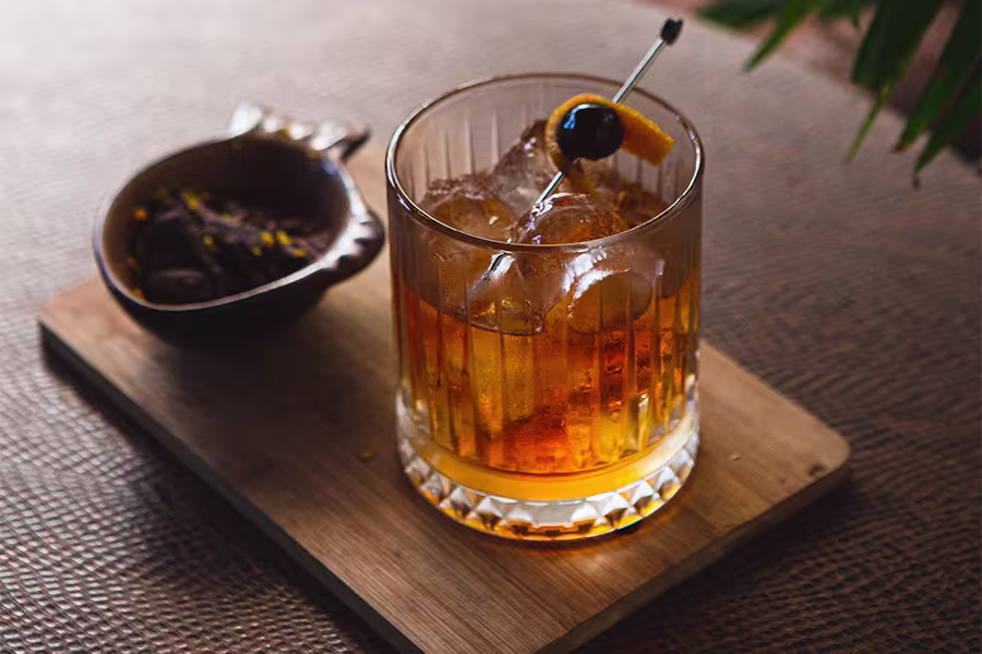 A glass of whiskey cocktail with ice, garnished with an orange peel and cherry, sits on a wooden board next to a small bowl of dried fruit and nuts.