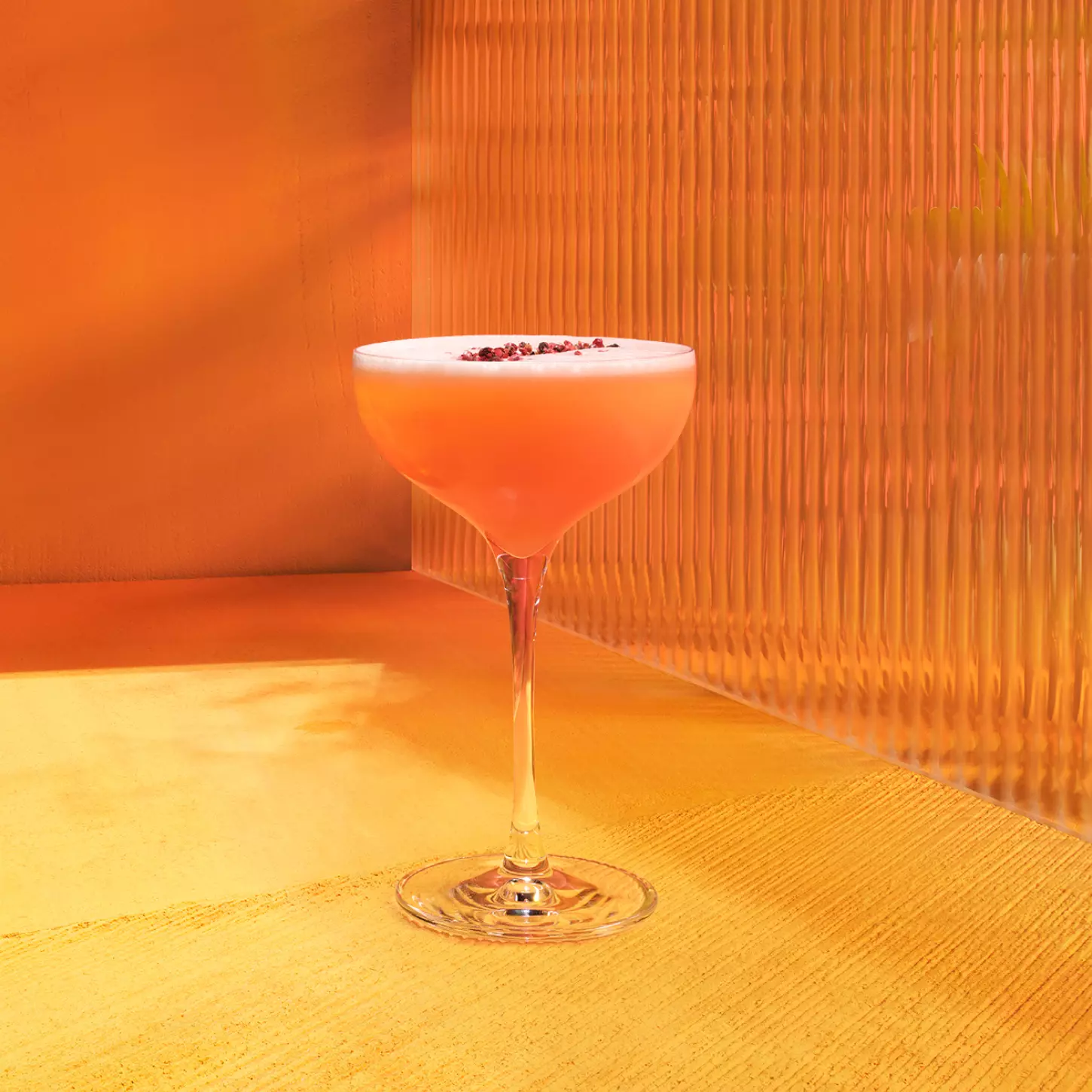 A coupe glass filled with an orange-pink cocktail topped with small garnish, set on a yellow and orange textured surface with a corrugated background.
