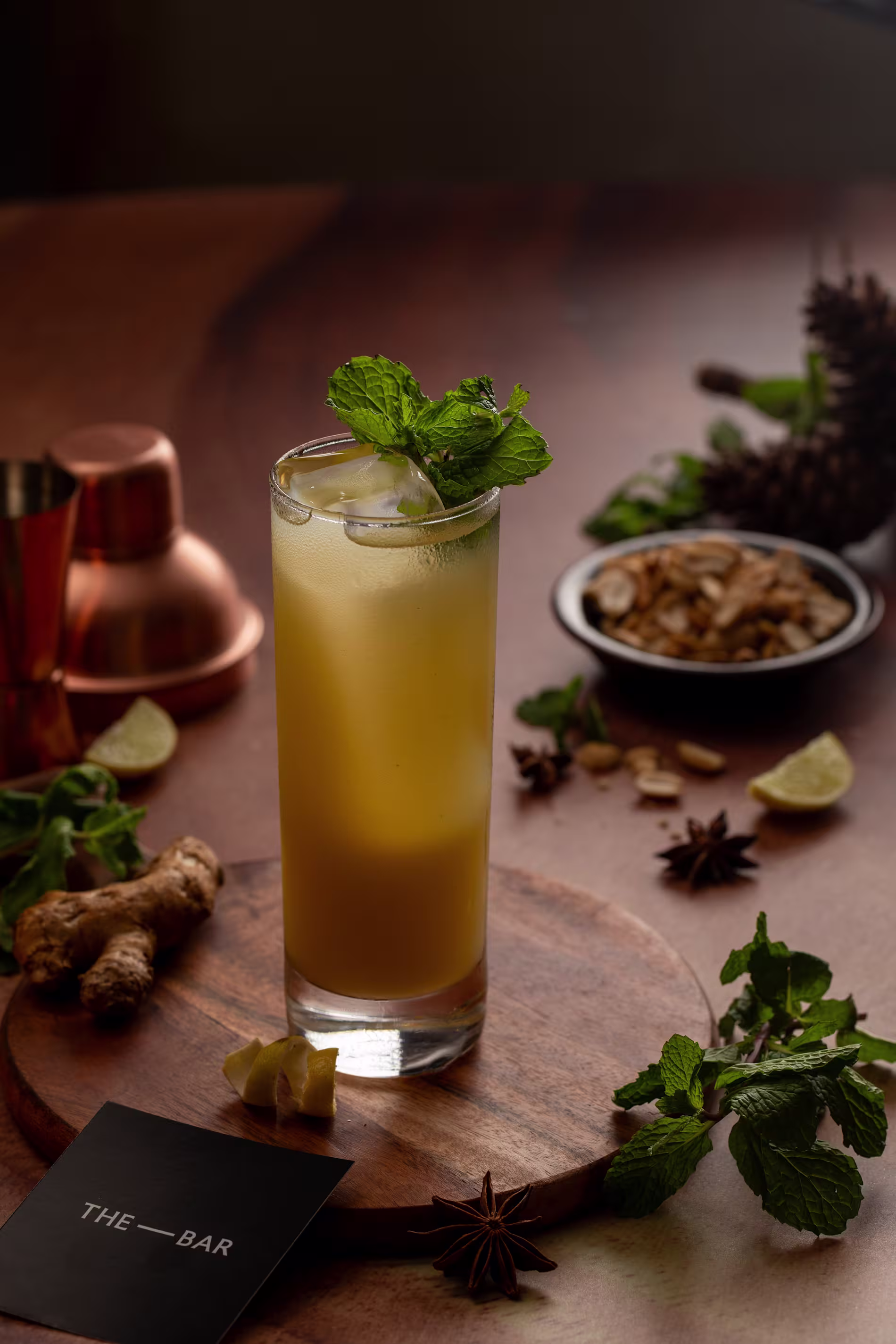 A tall glass of iced yellow drink garnished with mint, placed on a wooden board with ginger, lime, spices, mint leaves, and a bar card nearby.