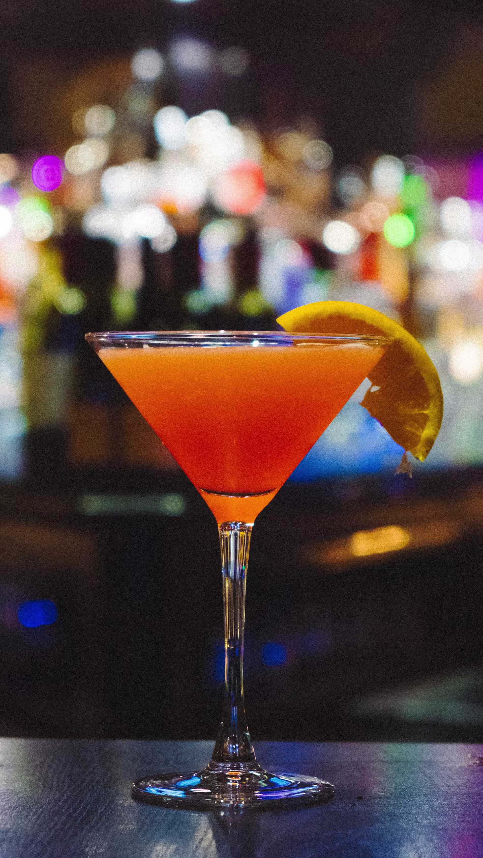 A martini glass filled with an orange-red cocktail, garnished with an orange slice, sits on a bar with blurred lights in the background.