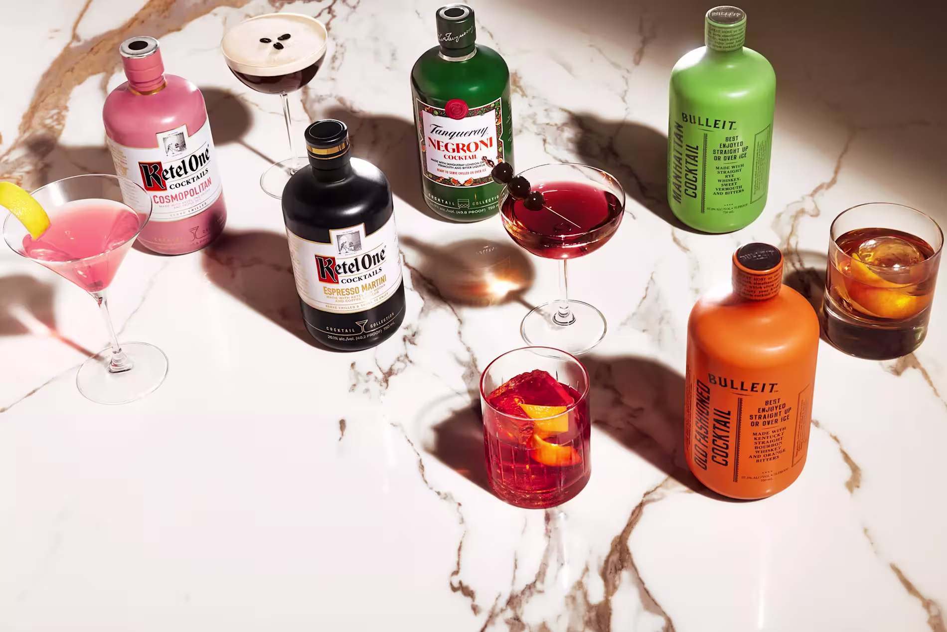 Various bottled cocktails and mixed drinks are arranged on a marble surface, each with a matching glass containing a garnish.