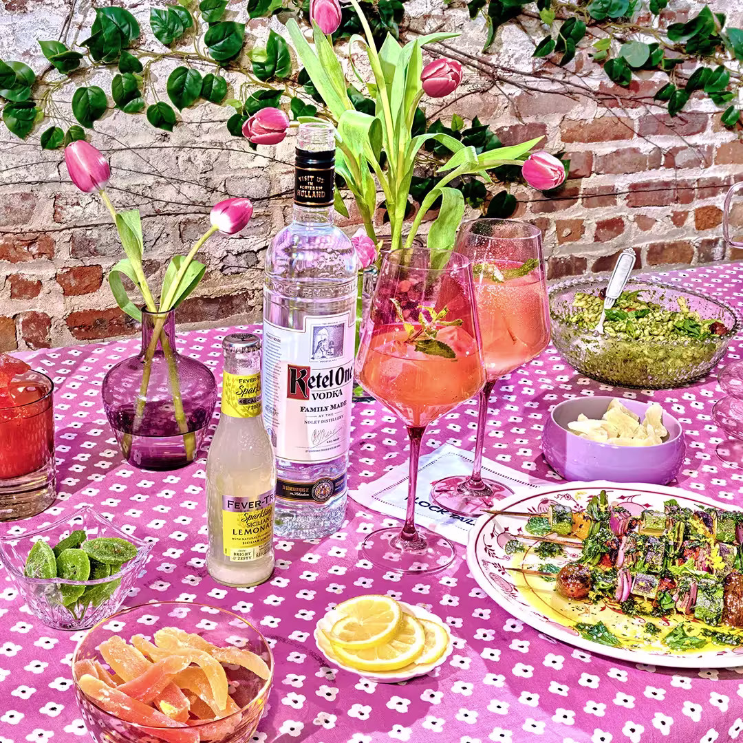 A pink-patterned table is set with vodka, tonic, cocktails, sliced citrus, mixed drinks, a salad, guacamole with chips, and vases of pink tulips, against a brick wall backdrop.