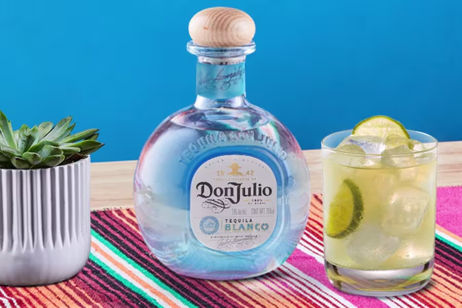 A bottle of Don Julio tequila sits on a colorful striped cloth next to a glass of cocktail with ice and lime, and a small potted succulent.
