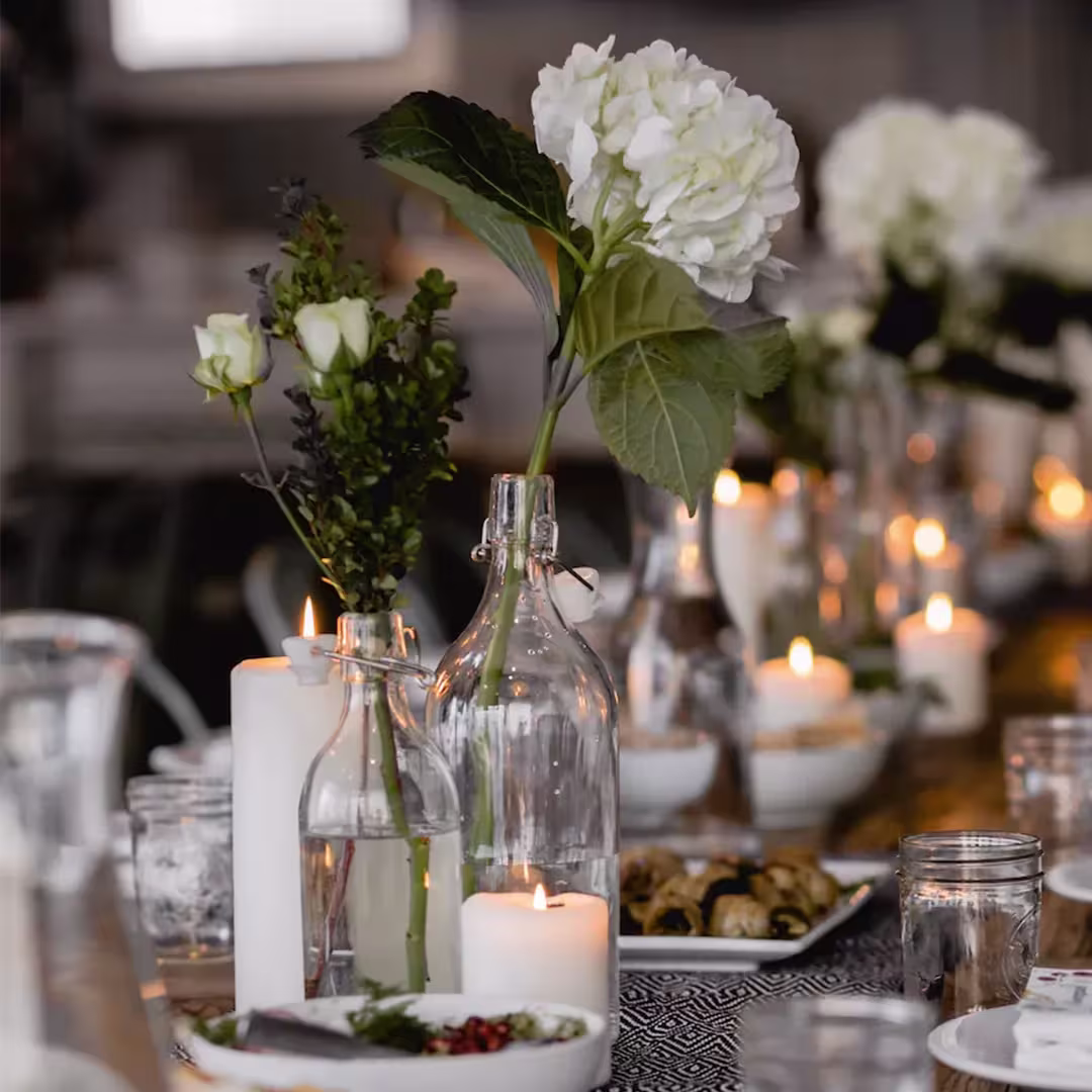 A dining table is set with glass vases holding white flowers, lit candles, bowls of food, and glassware, creating an elegant and cozy atmosphere.