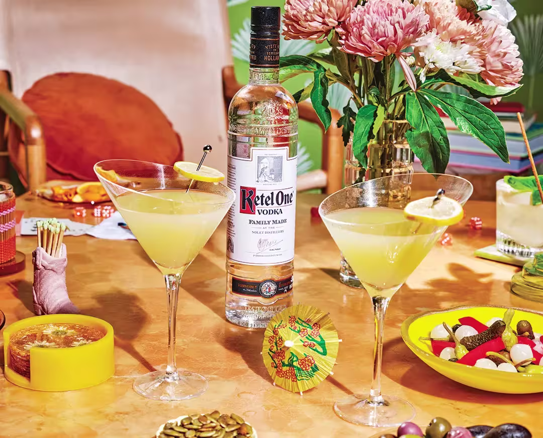 A bottle of Ketel One vodka is placed behind two martini glasses with yellow cocktails, surrounded by snacks, flowers, and assorted table items on an orange surface.