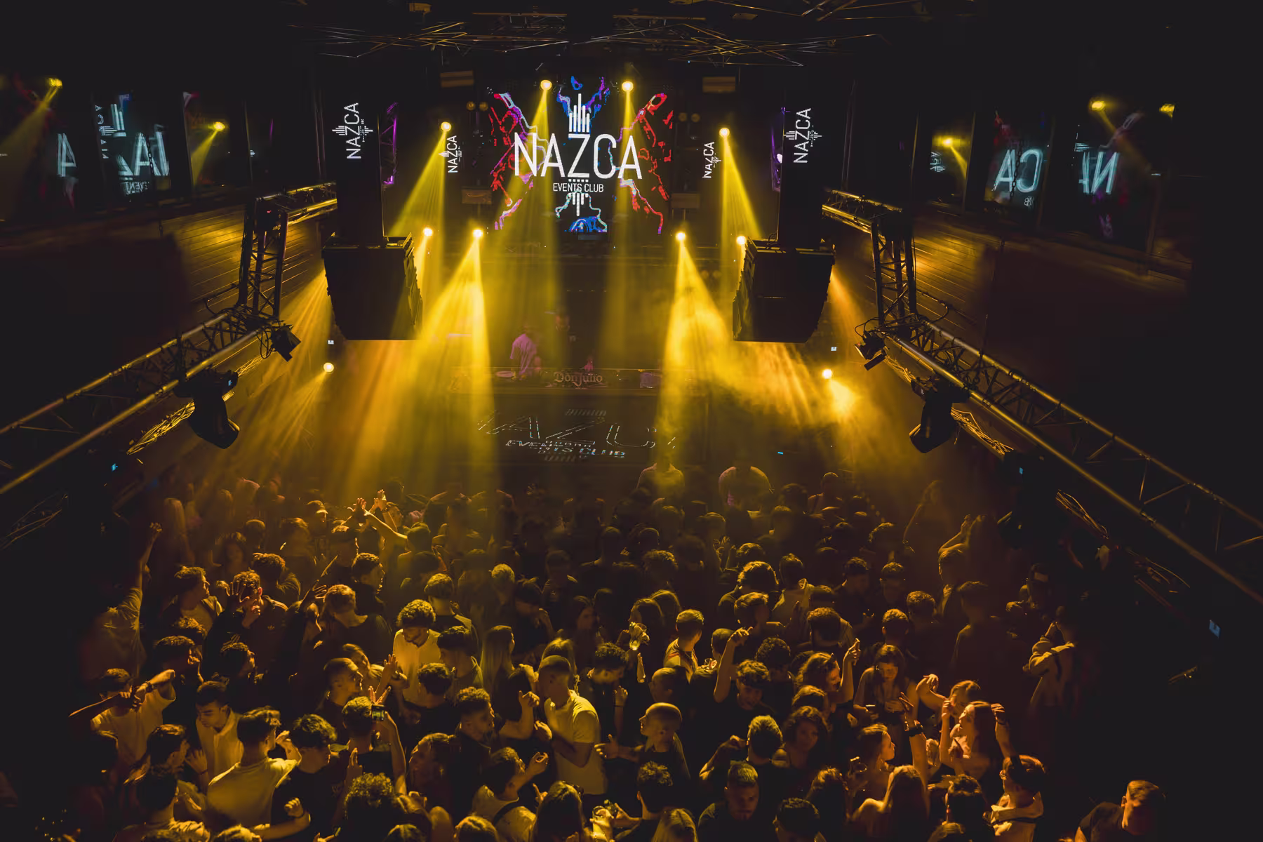 Pista llena en Nazca con luces doradas y DJ en el escenario. Ambiente vibrante y festivo en una noche de música electrónica.