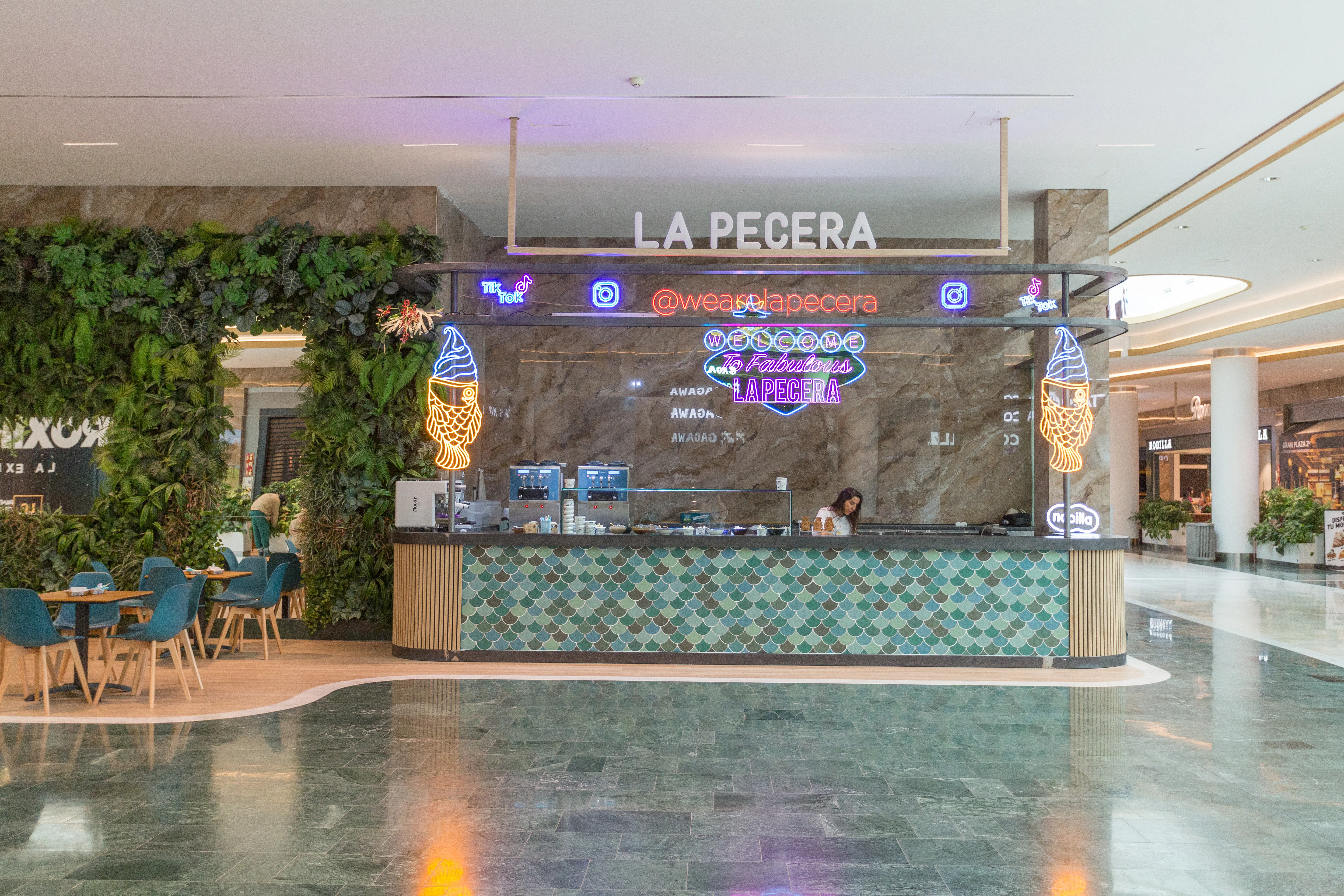 Mostrador de La Pecera con diseño de escamas, neones llamativos y una estética moderna que destaca entre vegetación y mármol.