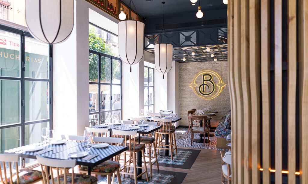 La foto muestra el interior de un restaurante Casa Berrinche. Hay varias mesas de comedor preparadas con platos, vasos y servilletas, listas para los comensales. El restaurante cuenta con grandes ventanas que permiten que la luz natural llene el espacio.