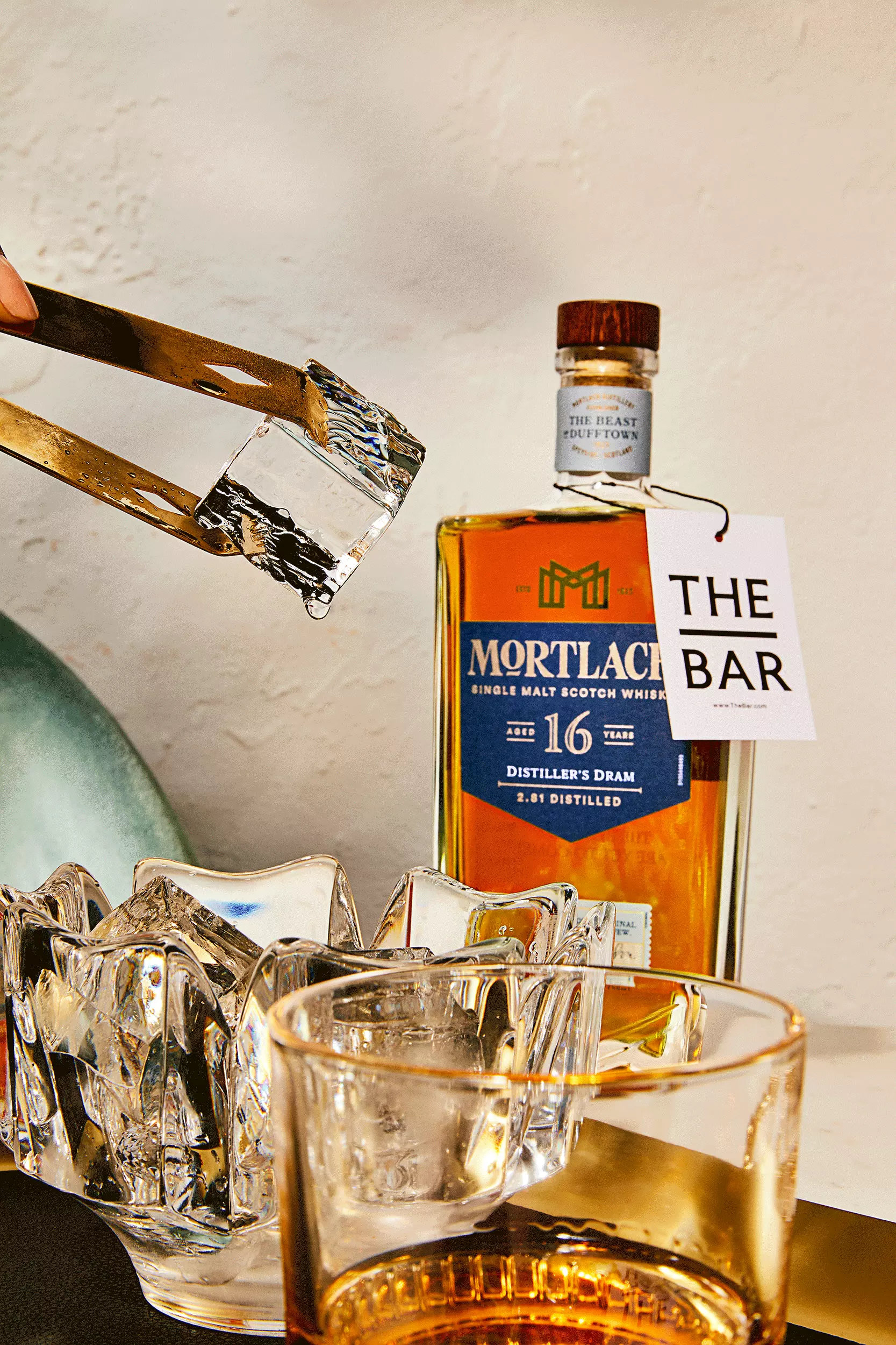 A hand uses tongs to place an ice cube into a glass, with a bottle of Mortlach 11-year-old single malt Scotch whisky and a THE BAR tag in the background.