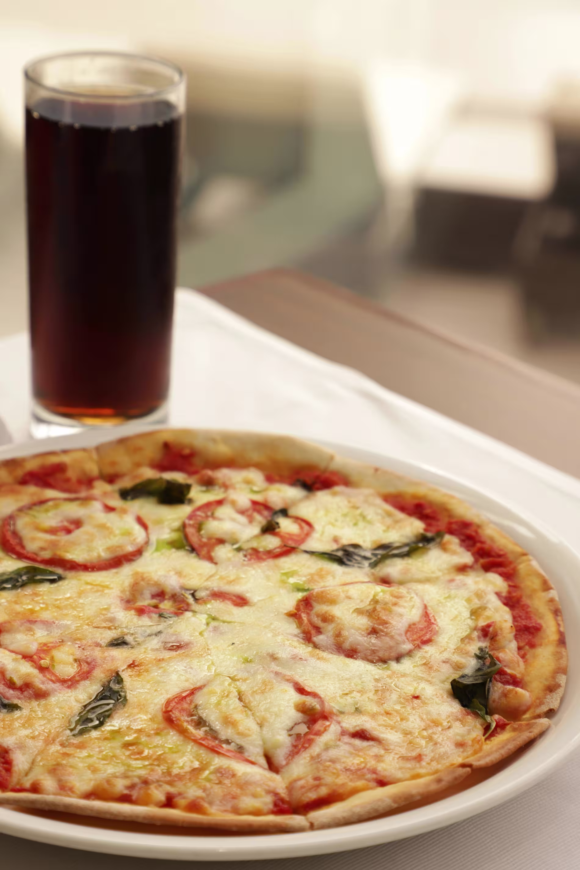 A cheese and tomato pizza on a white plate sits next to a glass of dark soda on a table.