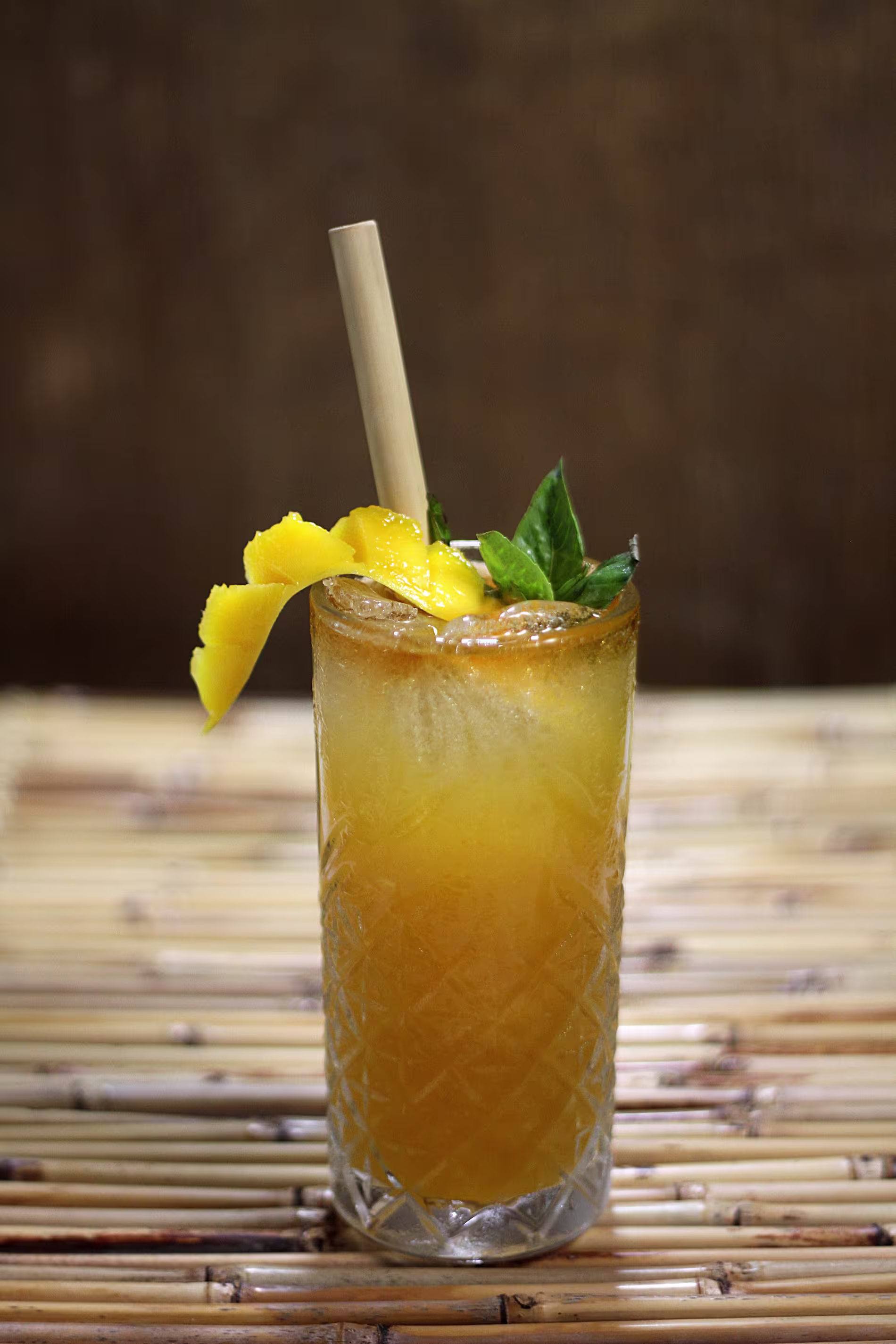 A tall glass filled with an orange cocktail, garnished with a twist of lemon peel and fresh mint, served with a straw on a bamboo surface.