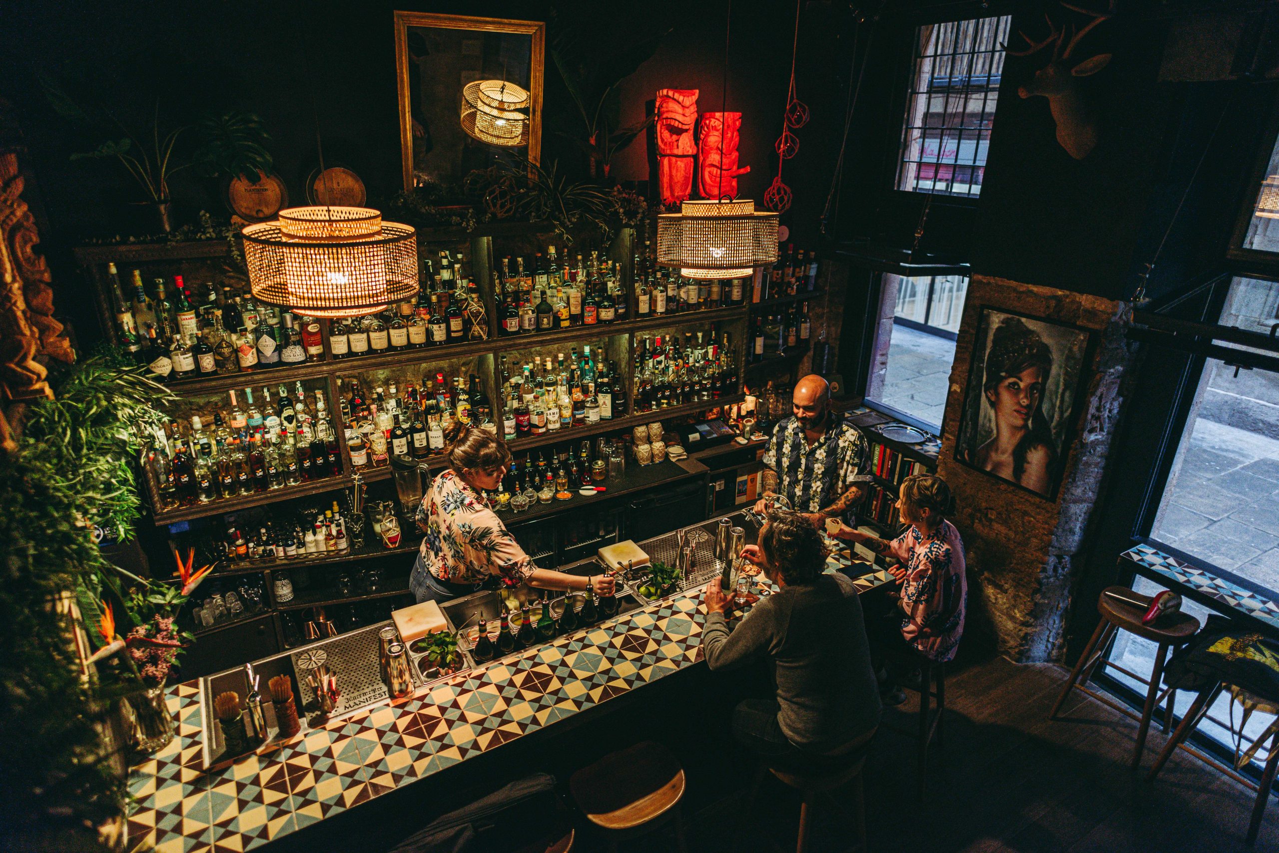 En Collage Bar, una barra de mosaicos vibrantes rodea estanterías repletas de botellas, plantas decorativas y arte en un ambiente cálido e íntimo.
