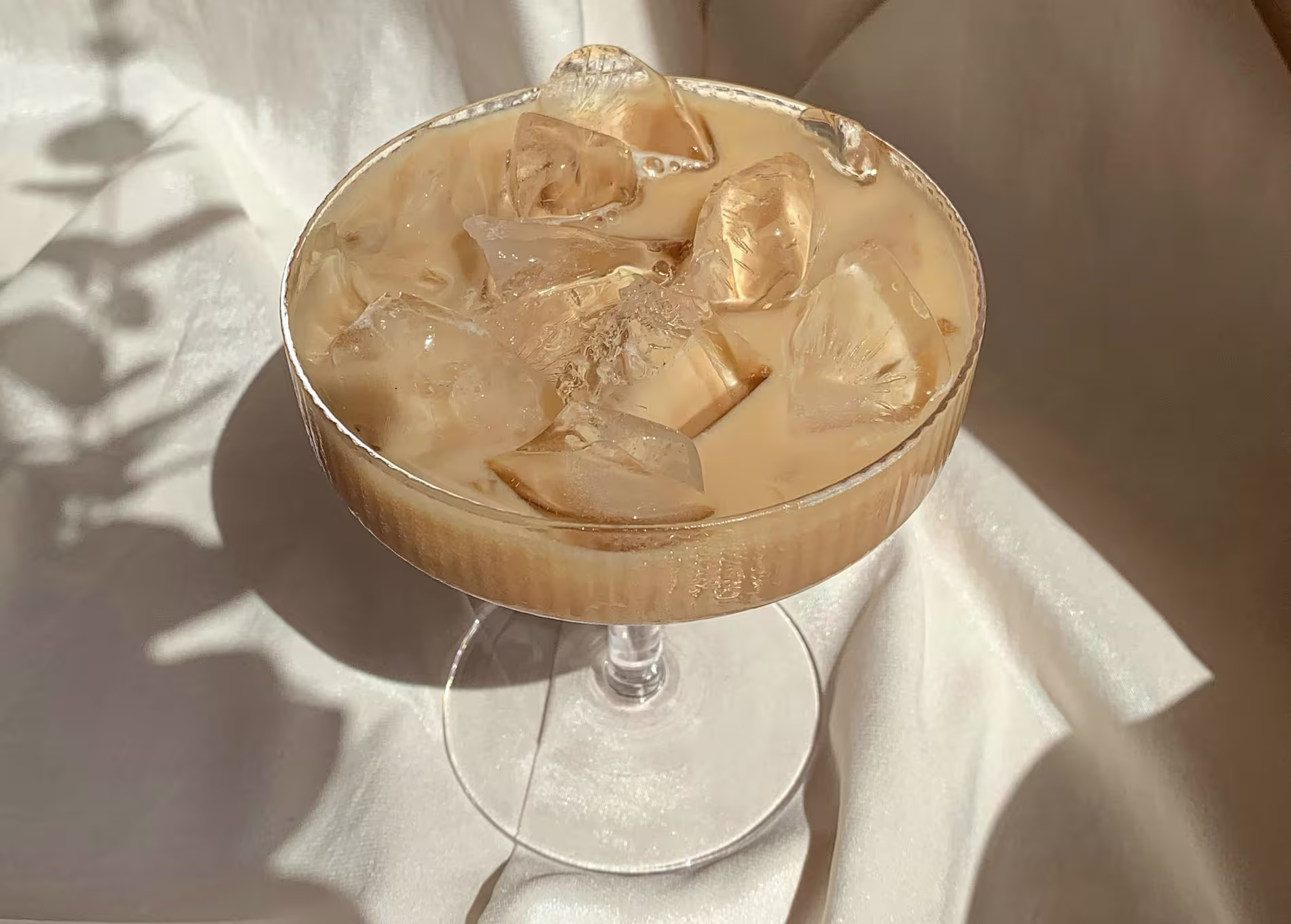 A coupe glass filled with iced coffee or a creamy iced beverage sits on a light satin fabric background.