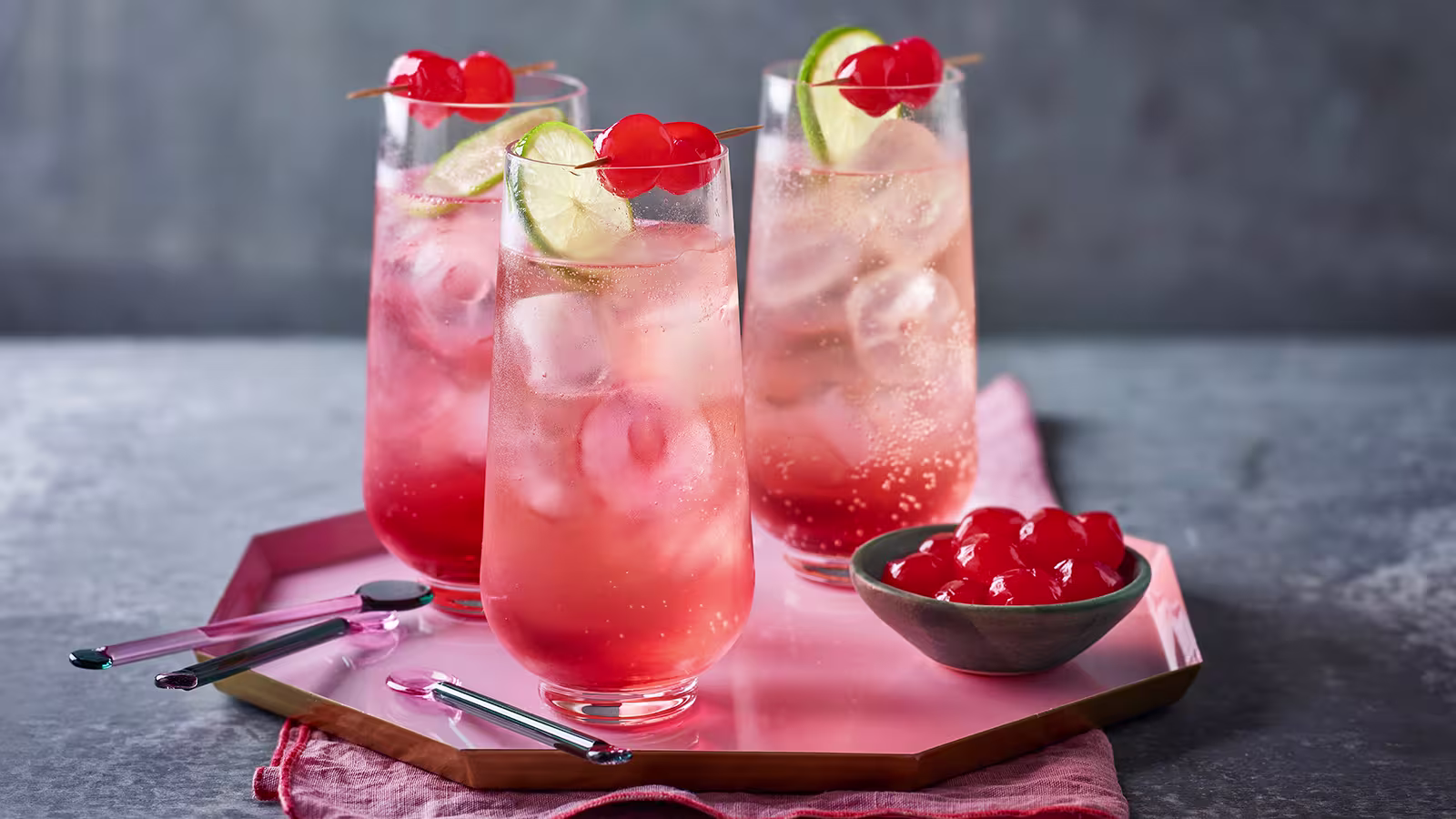 Three tall glasses of pink fizzy drink with ice, lime slices, and maraschino cherries, placed on a pink tray with a bowl of cherries and cocktail stirrers nearby.