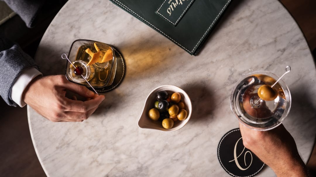 Dos cócteles servidos con aceitunas y piel de cítrico sobre mesa de mármol en Citrus Cocktail Bar by Sargis, junto a un cuenco de aceitunas y carta verde.