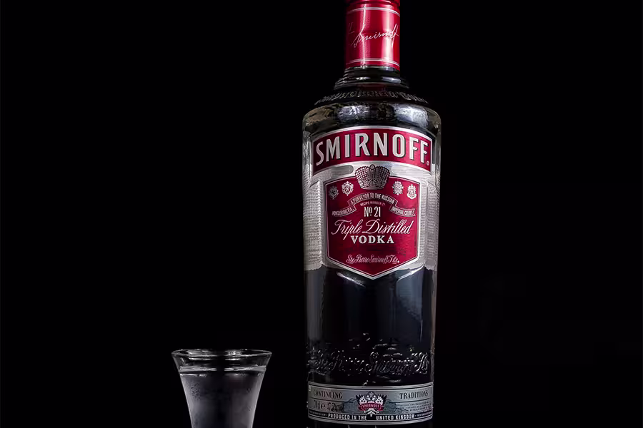 A bottle of Smirnoff No. 21 vodka stands next to a filled shot glass against a black background.