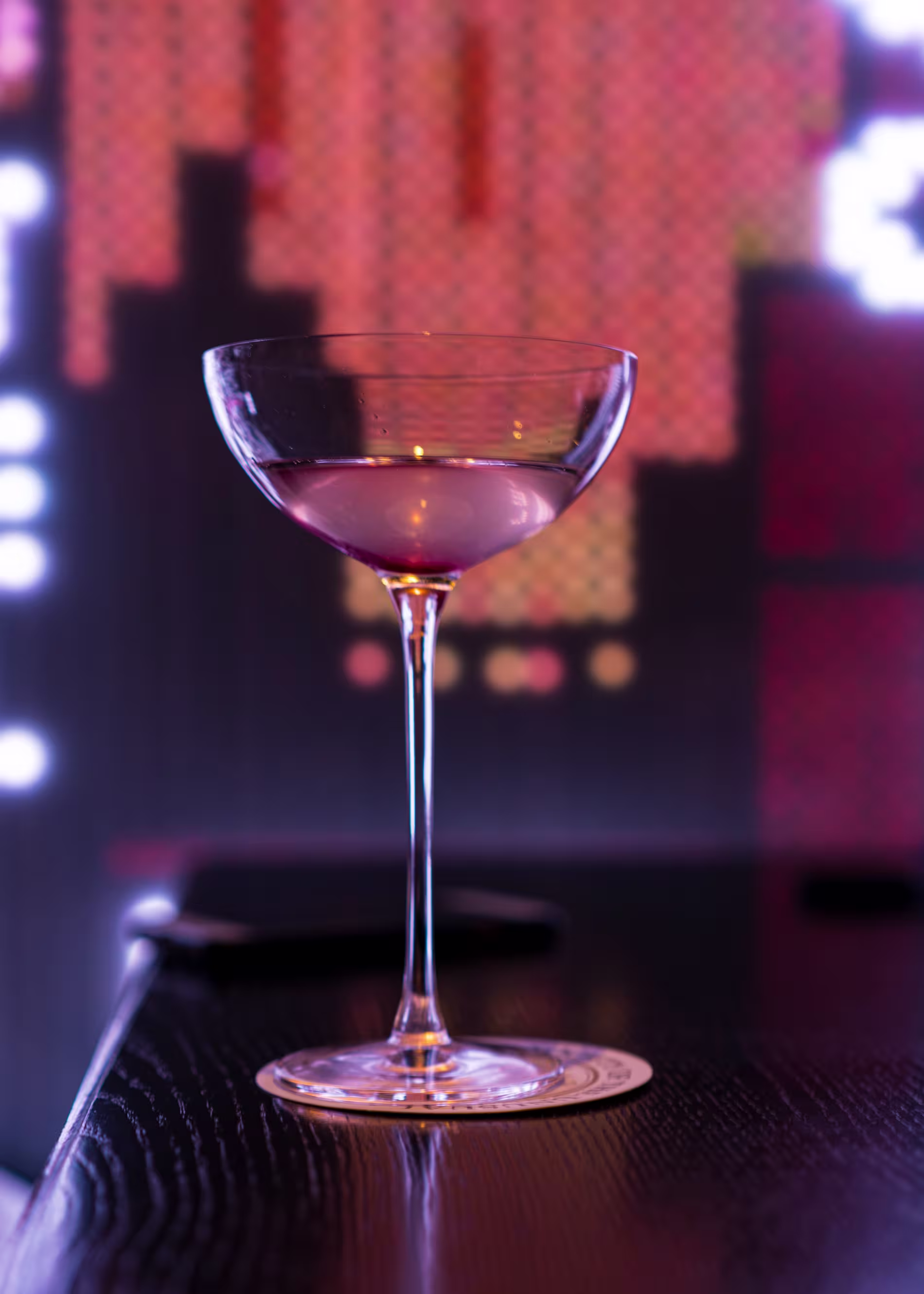 A coupe glass with a pink cocktail sits on a coaster atop a dark bar, with a blurry, colorful background of lights.