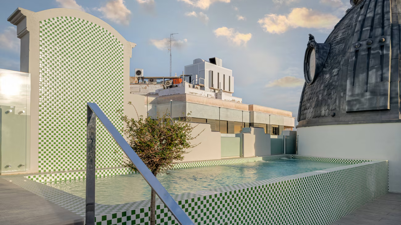 Piscina elevada con mosaico verde y blanco en una azotea urbana, rodeada de arquitectura clásica y moderna, bajo un cielo parcialmente nublado al atardecer.