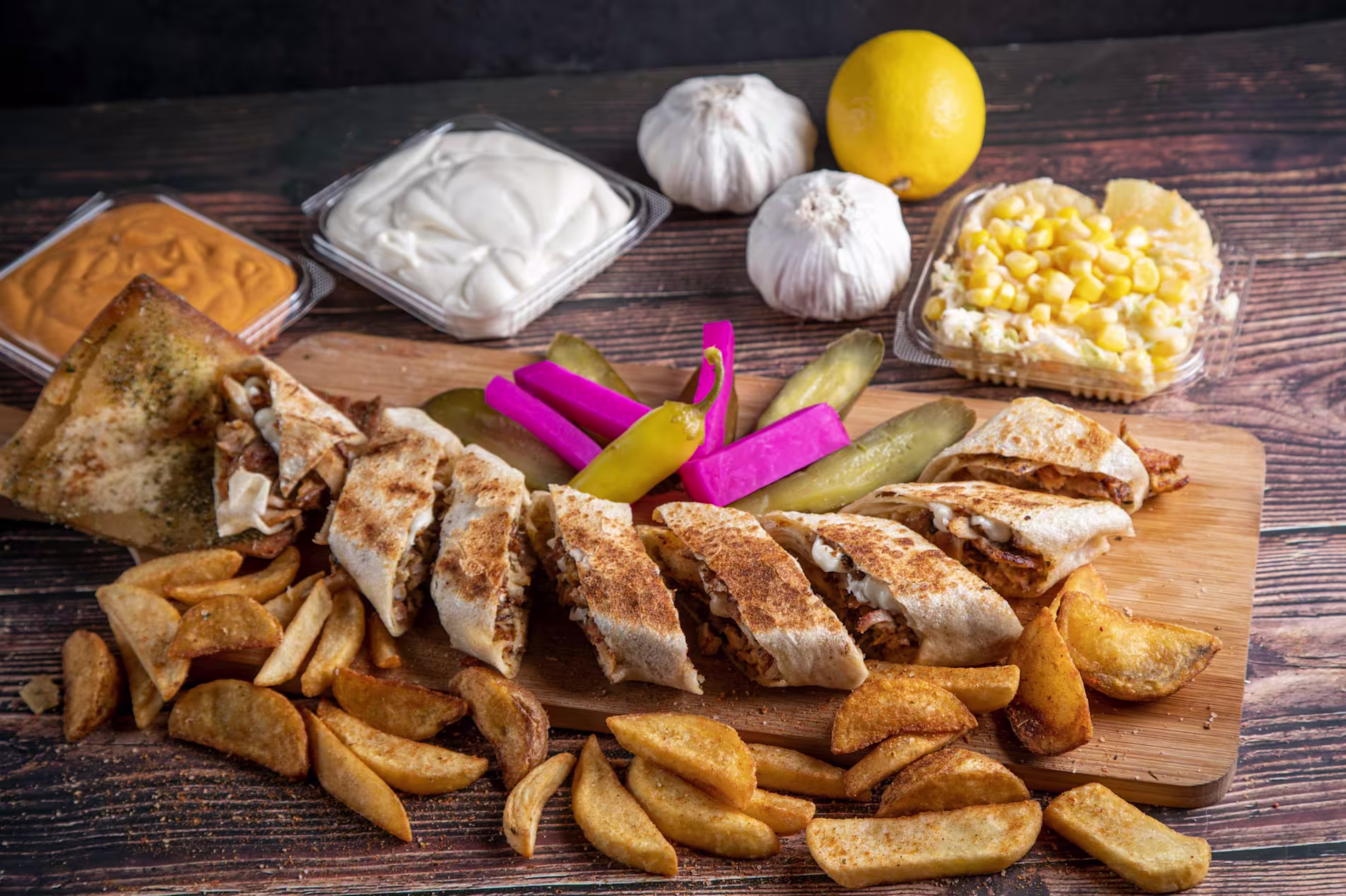 A sliced wrap with grilled filling is served with thick-cut fries, pickles, pickled turnips, sauces, corn, lemon, and garlic on a wooden board.