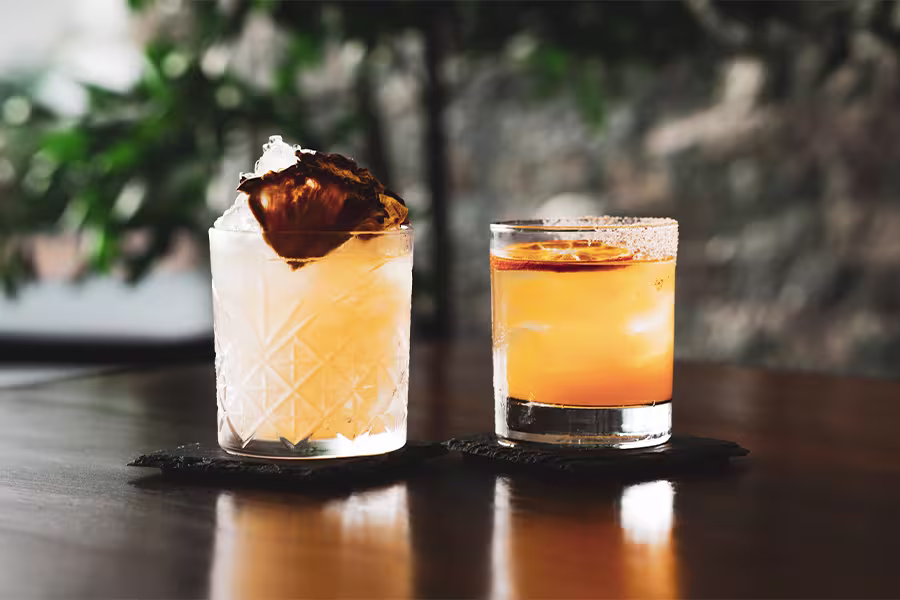 Two cocktails on black coasters sit on a wooden table; the left drink has ice and a dried garnish, the right is orange with a citrus slice.