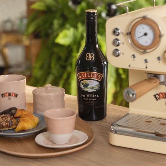 La imagen muestra una botella de Baileys Original Irish Cream, una cafetera y una bandeja que contiene dos tazas, una azucarera y un plato con alimentos dulces.