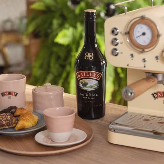 La imagen muestra una botella de Baileys Original Irish Cream, una cafetera y una bandeja que contiene dos tazas, una azucarera y un plato con alimentos dulces.