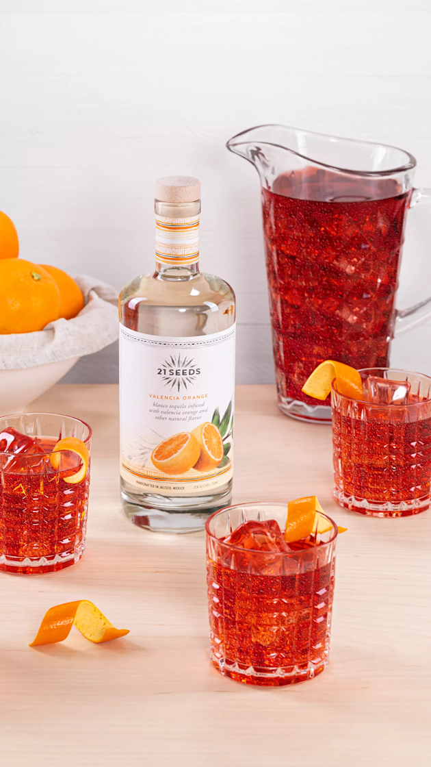 A bottle of 21Seeds Valencia Orange tequila, a pitcher, and three glasses of a red-orange cocktail with orange garnishes are arranged on a wooden surface near a bowl of oranges.