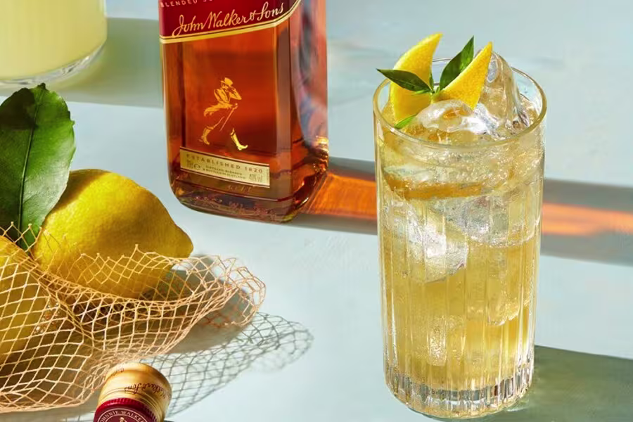 A glass of whisky with ice and lemon garnish sits beside a bottle of Johnnie Walker, lemons in a mesh bag, and a wine cork on a light blue surface.