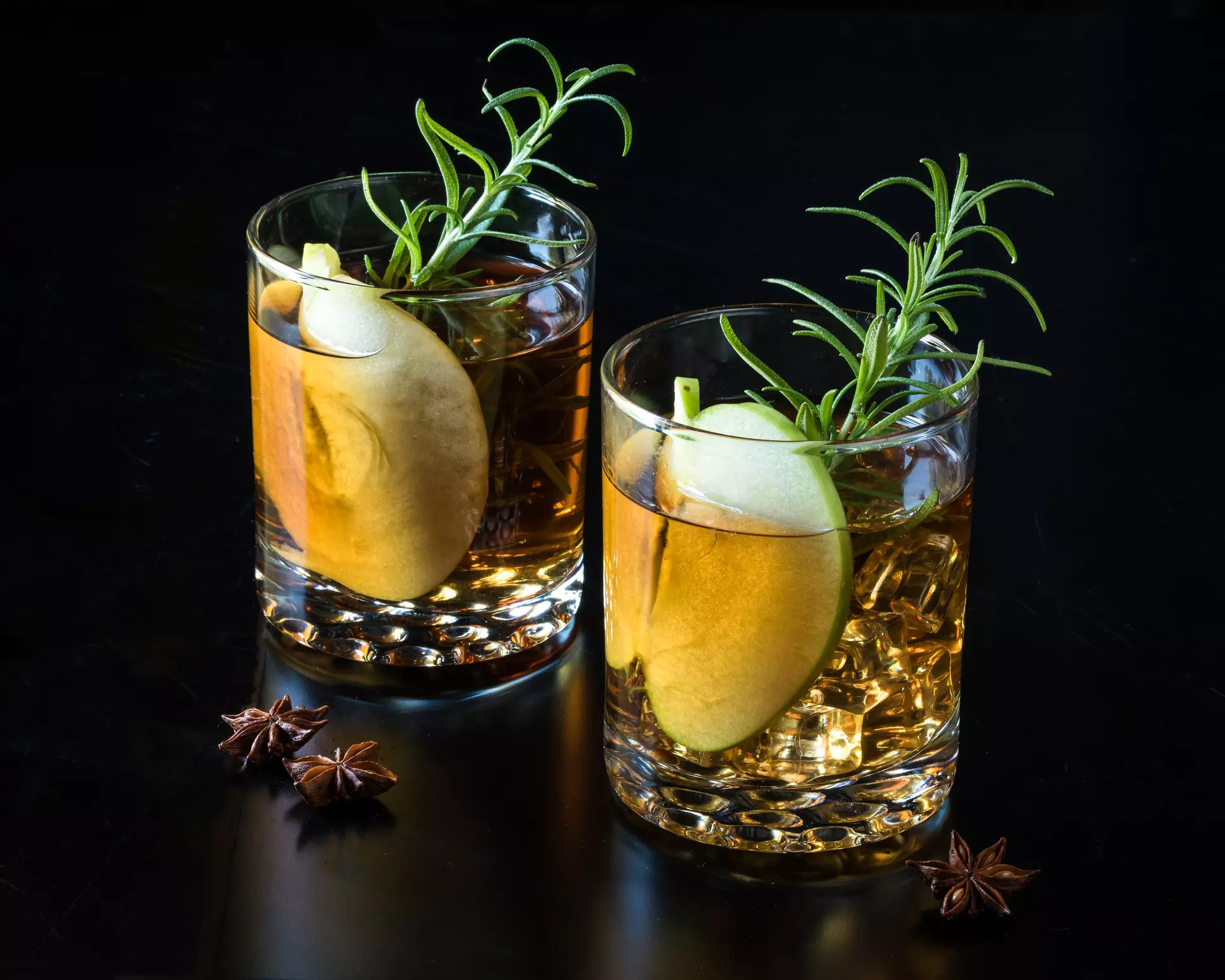Two glasses of amber-colored cocktails garnished with a green apple slice and a sprig of rosemary, with star anise scattered nearby on a black surface.