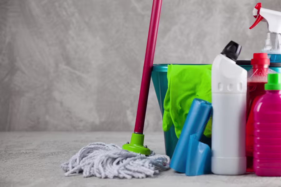 A set of cleaning supplies including a mop, bucket, gloves, spray bottle, trash bags, and various bottles of cleaners on a grey surface.