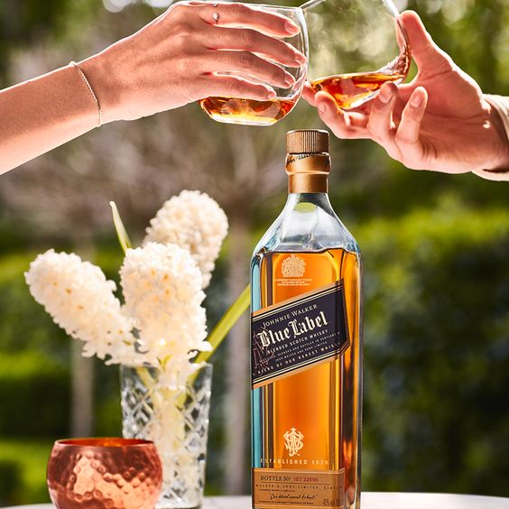 La imagen muestra una botella del whisky Johnnie Walker Blue Label, un brindis con dos vasos rellenos de dicho destilado y un florero con flores blancas.