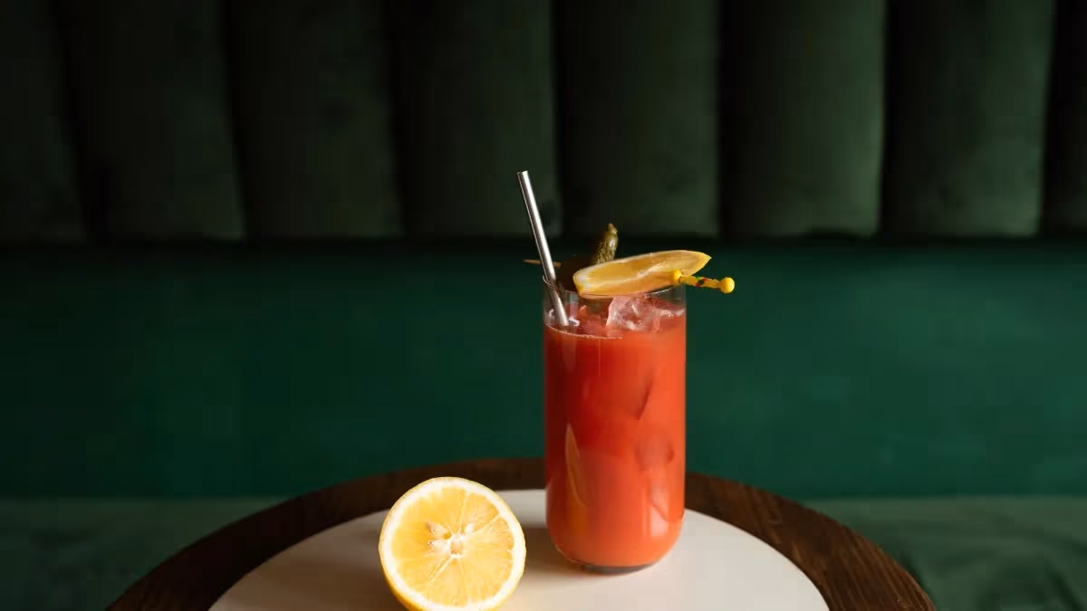 A tall glass of red cocktail with ice, garnished with a lemon slice and stir stick, sits on a table next to a halved lemon, with a dark green cushioned background.