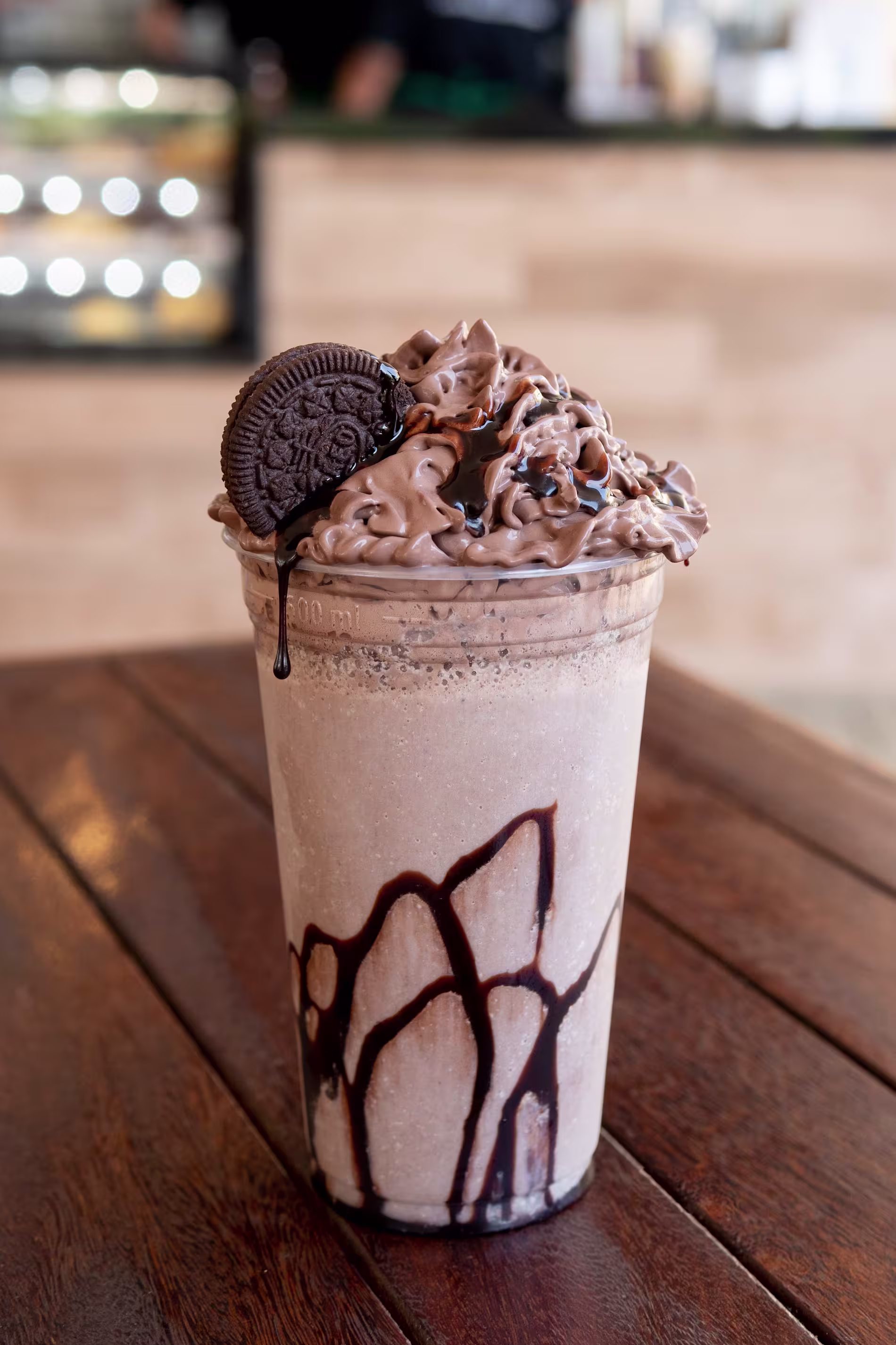 A chocolate milkshake in a clear plastic cup, topped with whipped cream, chocolate syrup, and a chocolate sandwich cookie, sits on a wooden table.
