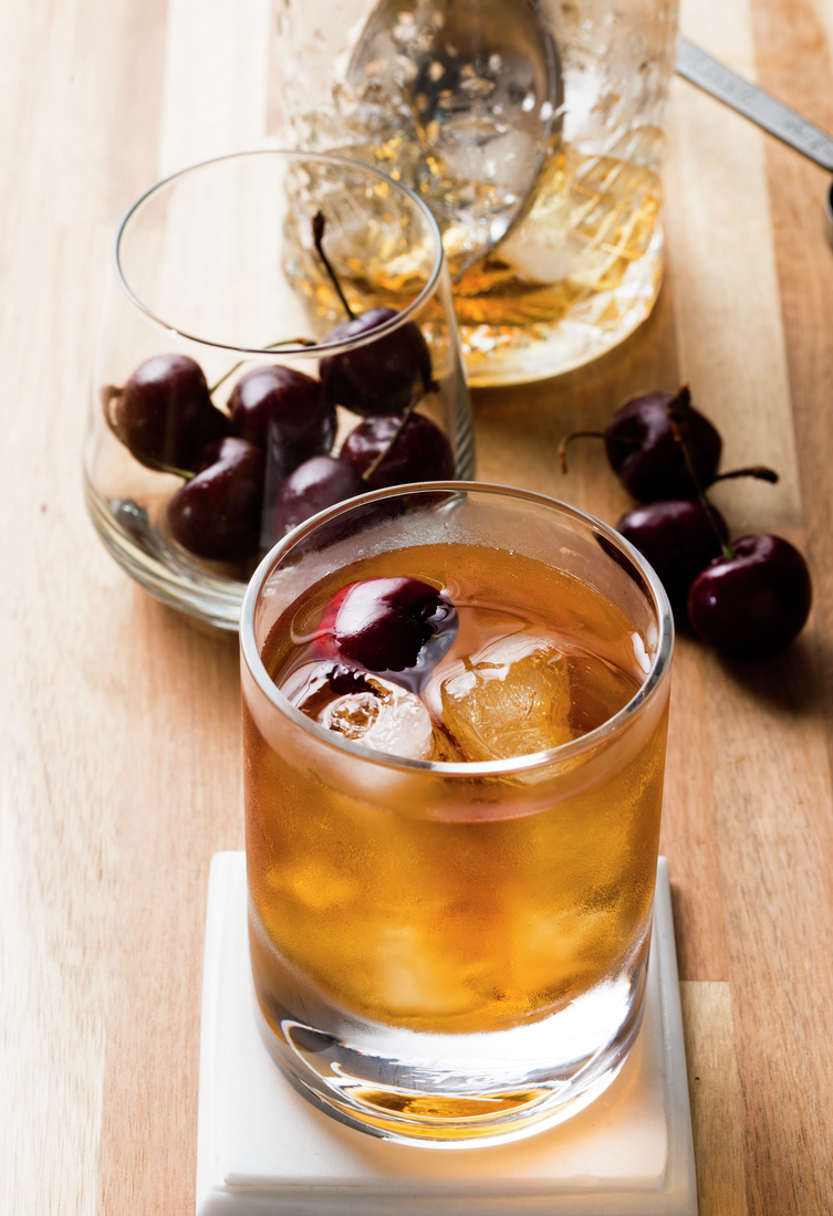 La imagen muestra un vaso relleno con el cóctel Manhattan, otro vaso relleno con cerezas y otros elementos de coctelería alrededor.