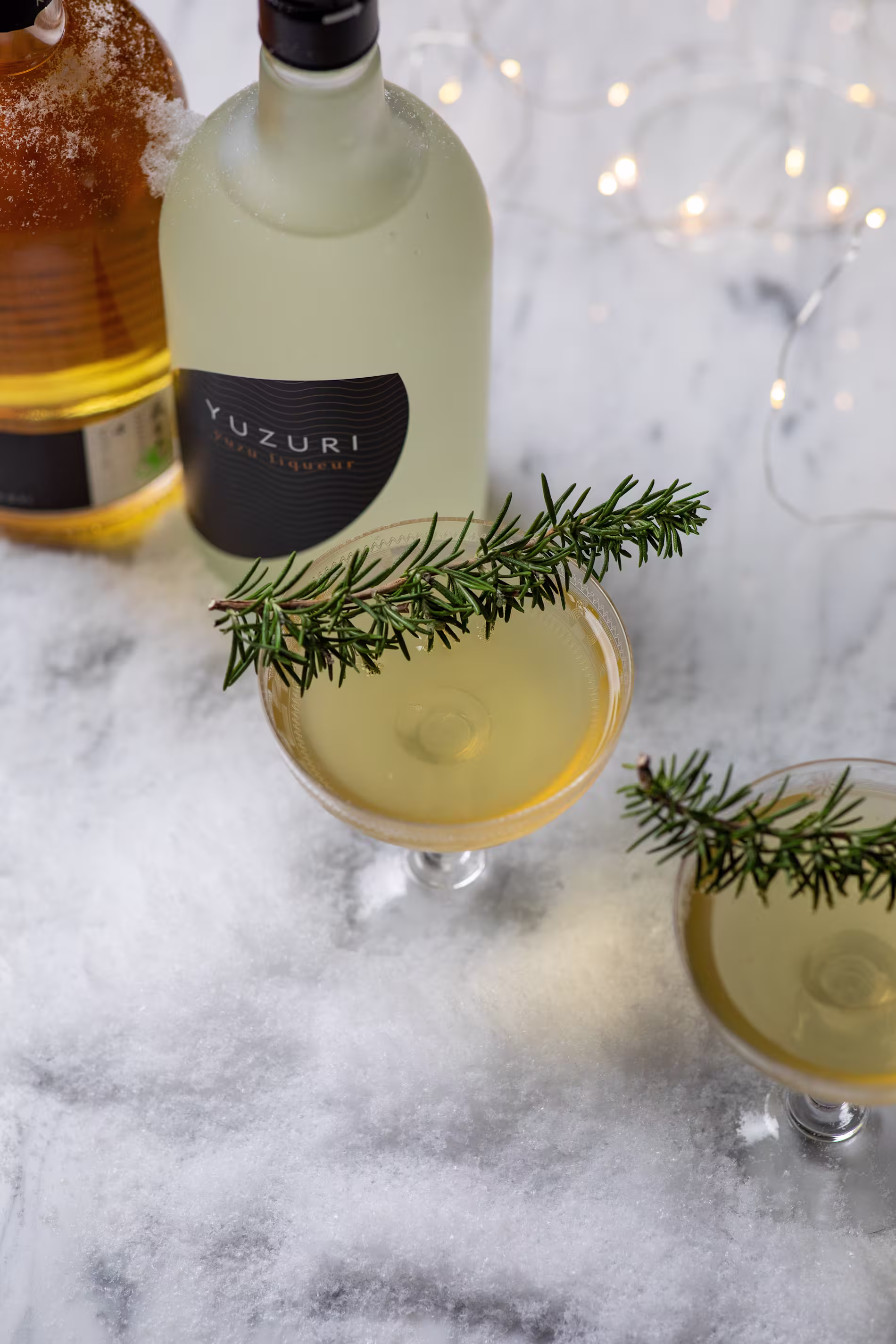Two cocktails garnished with rosemary sprigs sit on a snowy surface near two bottles, one labeled Yuzuri, with string lights in the background.