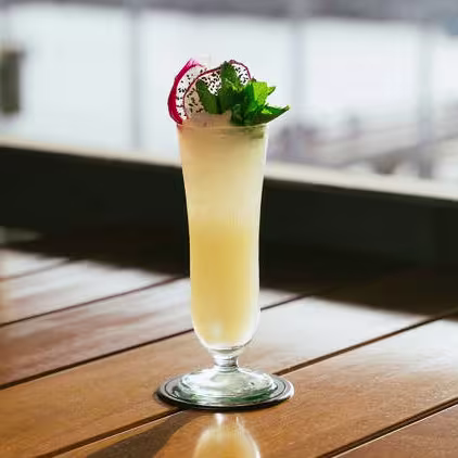 Cóctel tropical servido en copa alta, decorado con menta y pitahaya, sobre mesa de madera con vistas al puerto. Refrescante y elegante.