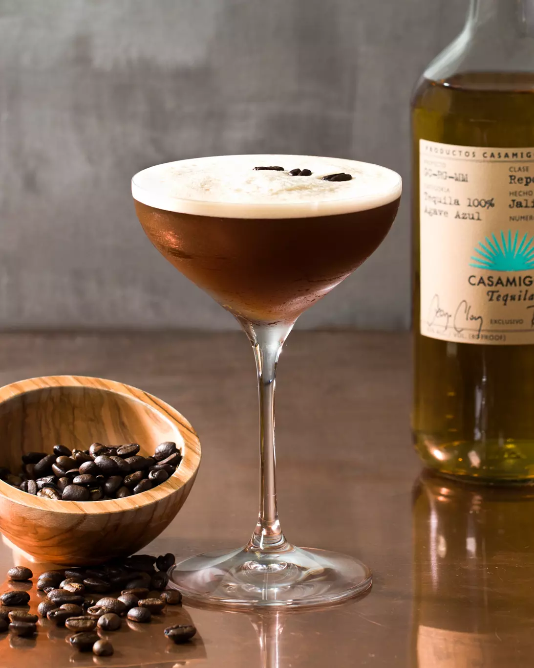 A cocktail in a coupe glass garnished with coffee beans, next to a wooden bowl of coffee beans and a bottle of Casamigos tequila.
