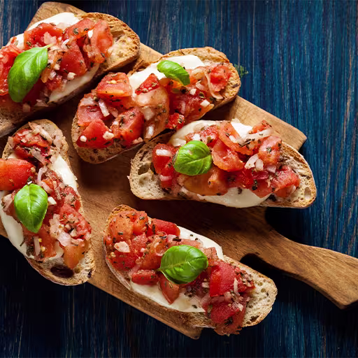 Slices of toasted bread topped with diced tomatoes, onions, chopped herbs, and fresh basil leaves are arranged on a wooden serving board.
