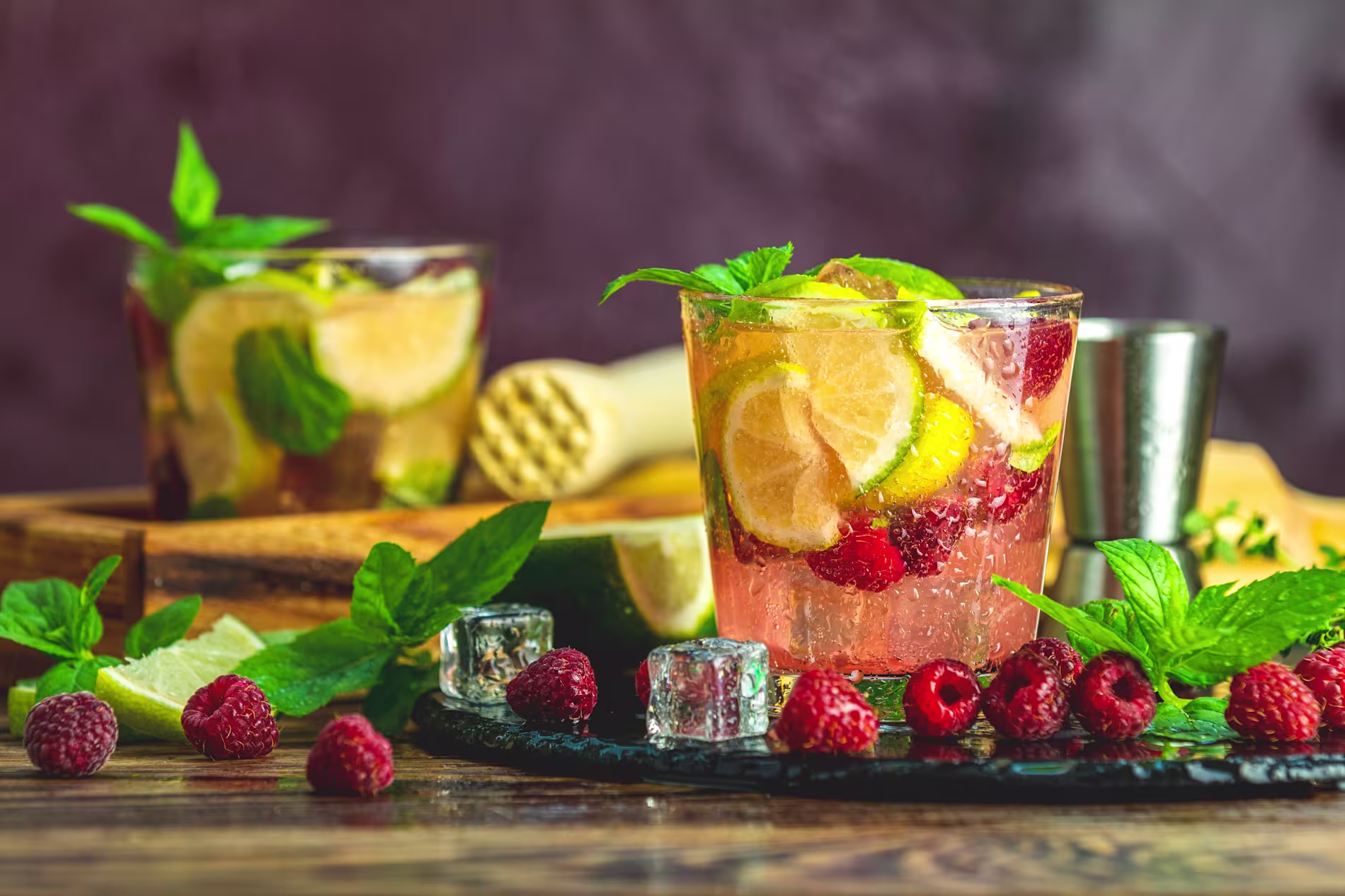 Two glasses of a fruit cocktail with lime, raspberries, mint leaves, and ice cubes on a wooden table; cocktail-making tools and ingredients in the background.