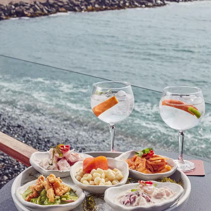 La imagen muestra un plato con 5 pequeños platos encima con diferentes comidas, dos copas llenas con mucho hielo, y el mar de fondo