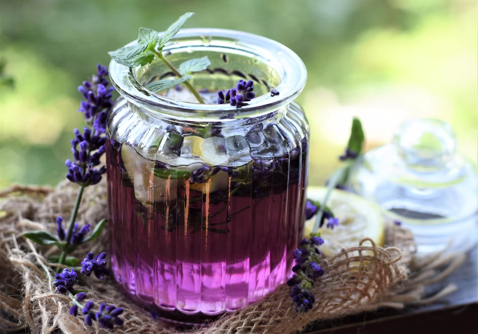 A glass jar filled with a purple drink, ice cubes, mint, and lavender sprigs, sits on burlap with lemon slices and additional lavender nearby.