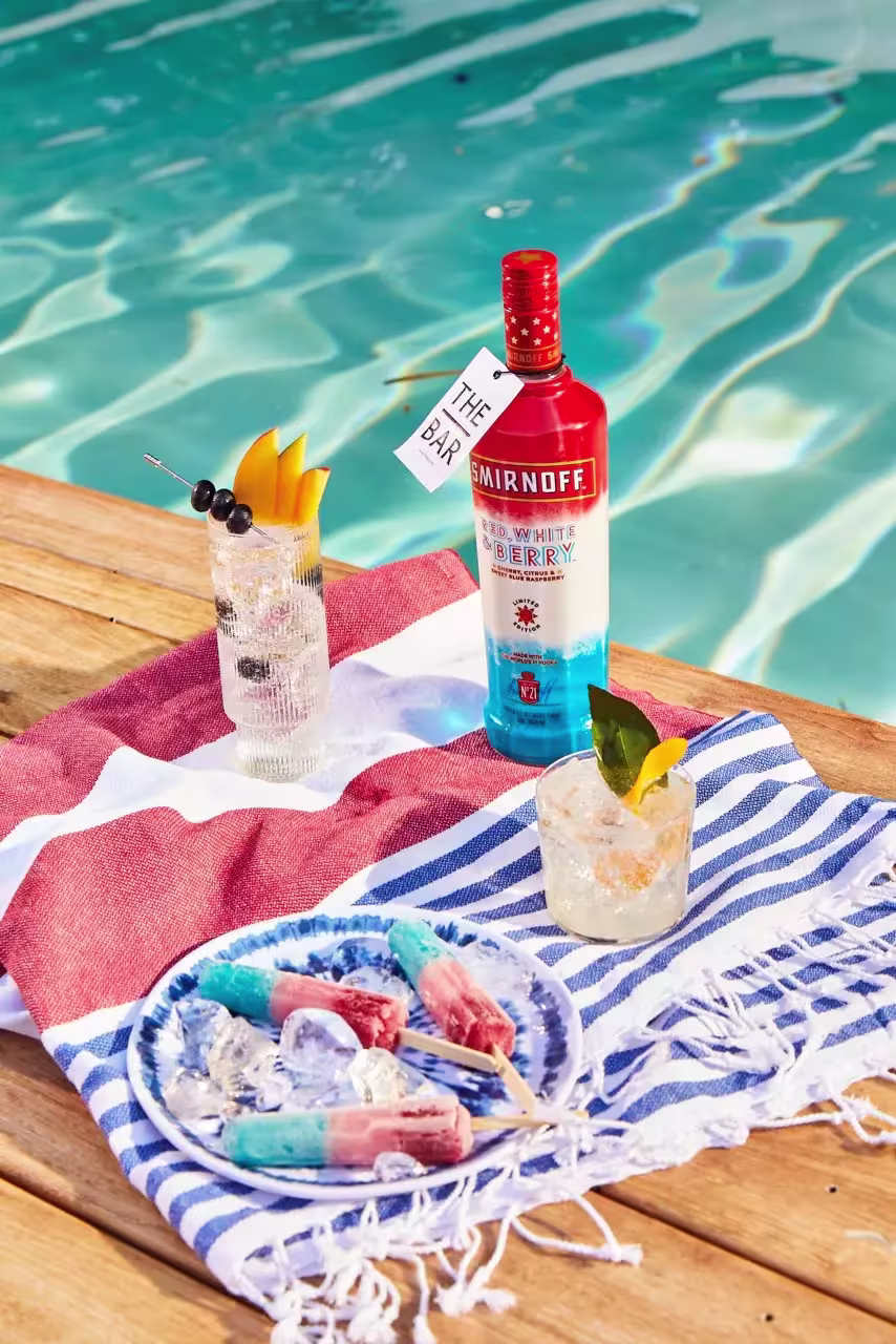 A bottle of Smirnoff Red, White & Berry vodka, three cocktails, and popsicles on a towel by a swimming pool.